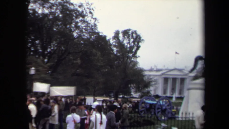 Protest crowd near the White House in Washington, DC, with fencing, display cannons, trees, and banners visible.