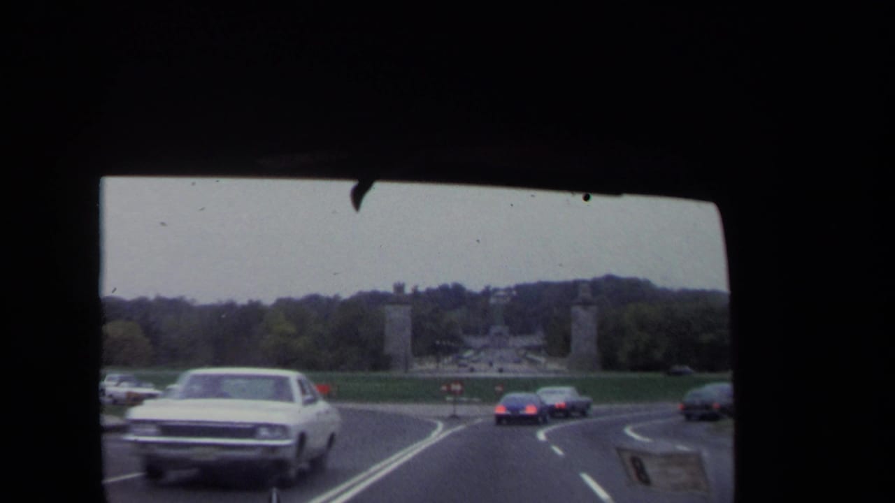 Cars travel on a divided road toward tall monument pylons and open lawns.