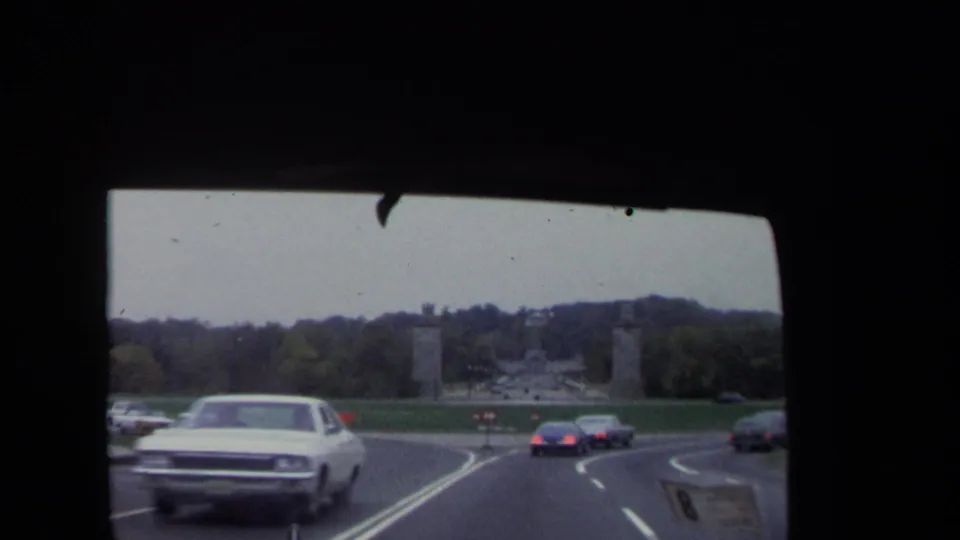 Moving-car view in Washington, DC showing 1970-era traffic, road markings, lawns, and distant monument pylons.