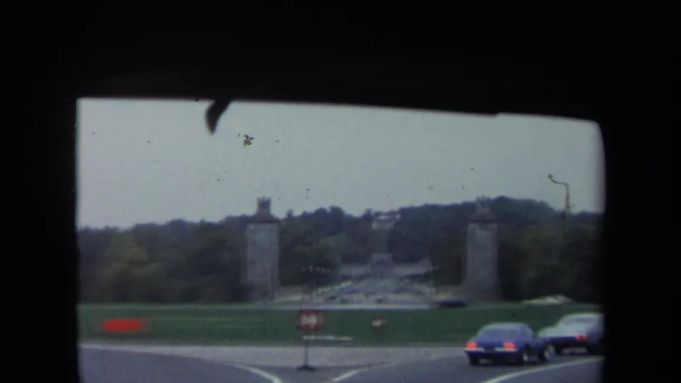 Washington, DC roadway frame showing lawns, monument pylons, passing cars, and red-and-white traffic direction signs.