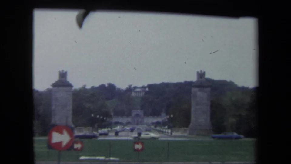 1970 Washington, DC home-movie frame looking across a roadway and lawn toward monument pylons, traffic, and a red arrow sign.