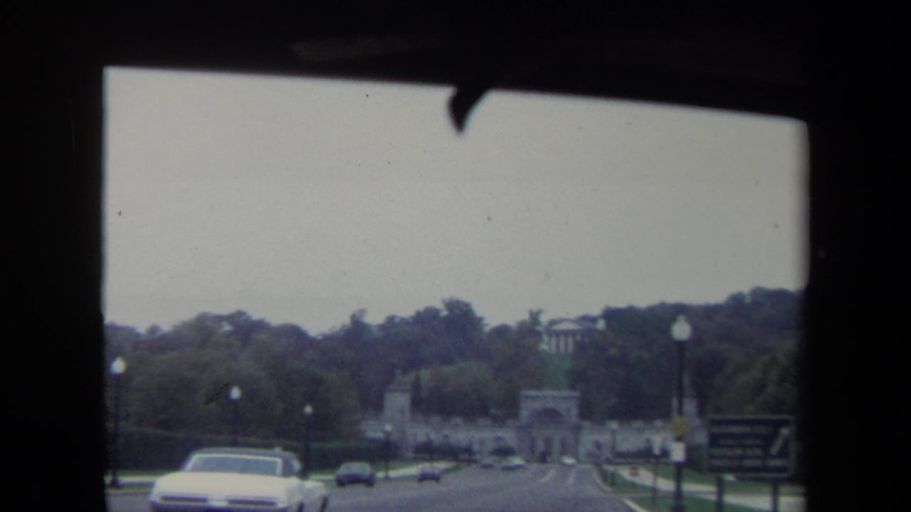 Roadway leading toward a stone archway, seen through a car windshield with vehicles and trees ahead.