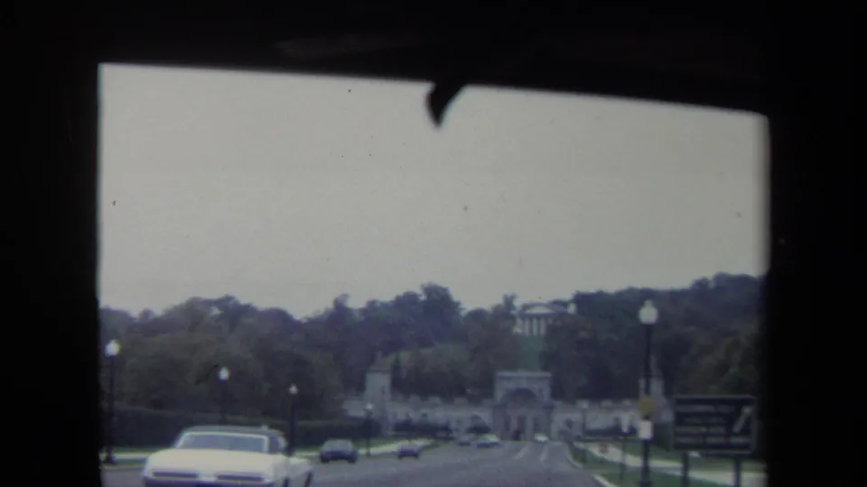 1970 Washington DC road approach toward a monumental stone archway, filmed from inside a moving car with traffic and trees visible.