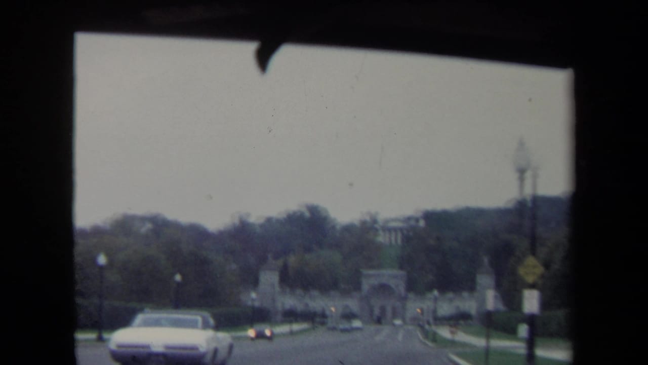 Washington DC driving view from 1970 with a white period automobile passing before a distant stone archway and tree-lined grounds.