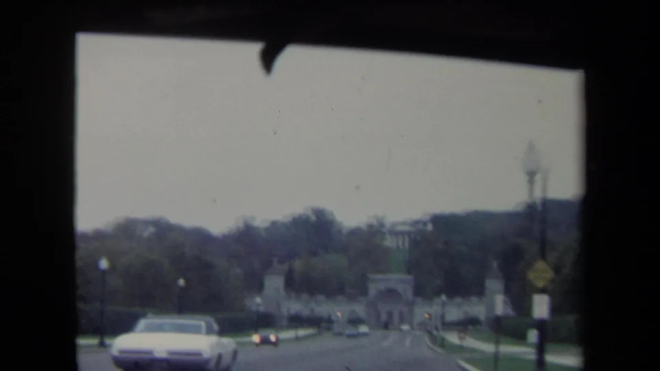 Washington DC driving view from 1970 with a white period automobile passing before a distant stone archway and tree-lined grounds.
