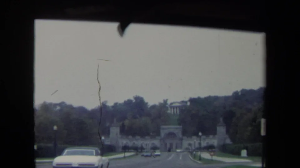 Soft 8mm frame of a Washington DC road approach, with a period car, tree-lined sides, and a stone gateway ahead.
