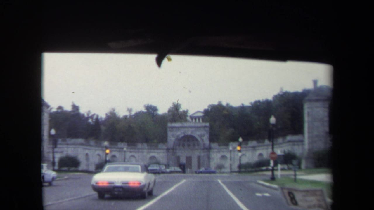 White sedan drives toward a large stone archway entrance on a tree-lined road.