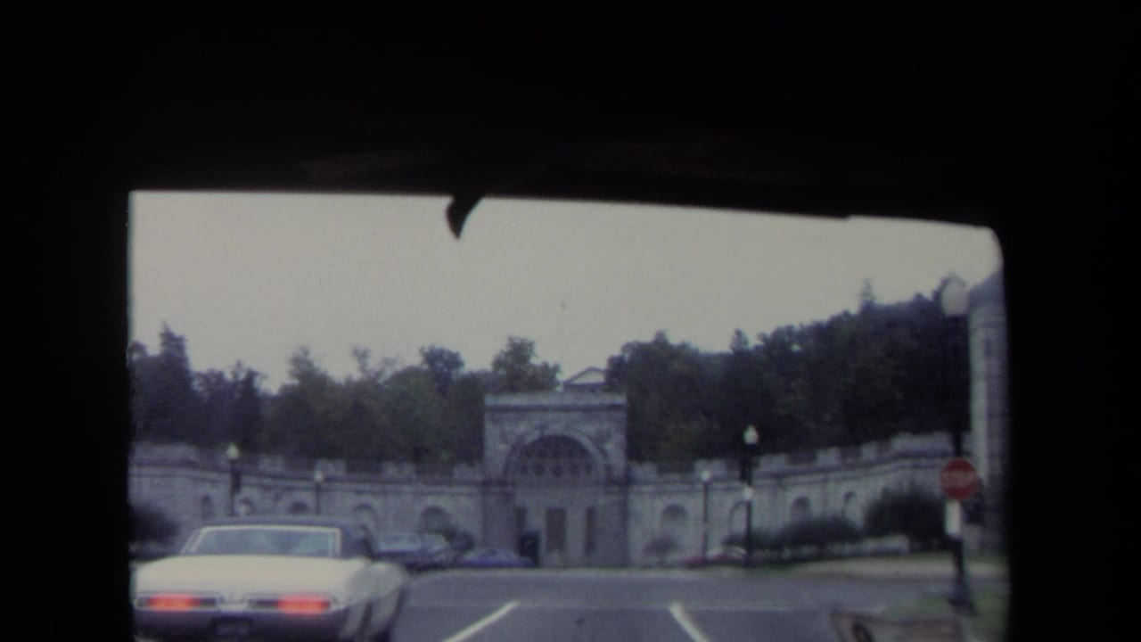1970 Washington, DC home-movie frame with a white car in the foreground and a large arched stone gateway set against trees and roadside signs.