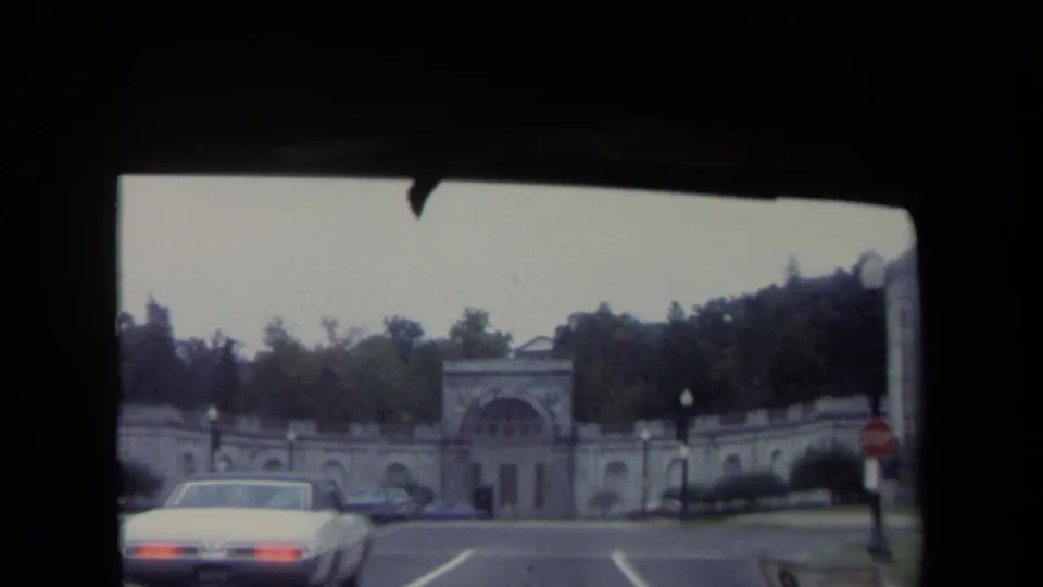 1970 Washington, DC home-movie frame with a white car in the foreground and a large arched stone gateway set against trees and roadside signs.