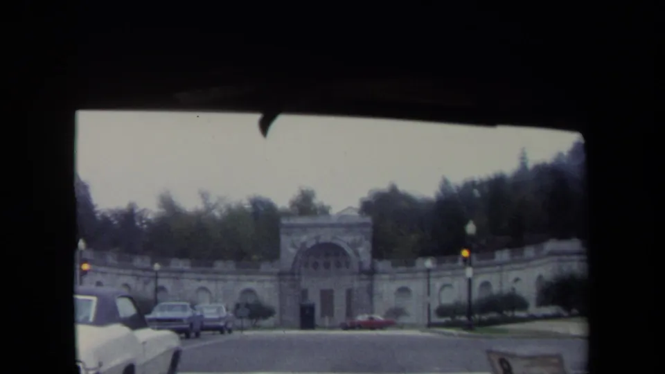 1970 Washington, DC 8mm frame from a moving vehicle showing a stone gateway, period cars, lamps, and a tree-covered rise beyond the entrance.