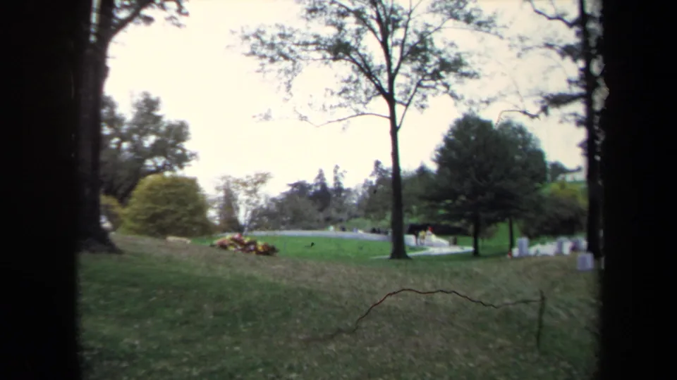 1970 Washington, DC 8mm frame showing a green cemetery-like park with mature trees, scattered gravestones, flower beds, and distant visitors.