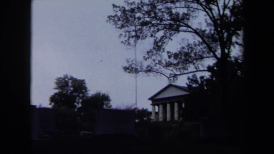 1970 8mm frame of a classical columned structure in Washington DC parkland, partly obscured by trees and shadow.