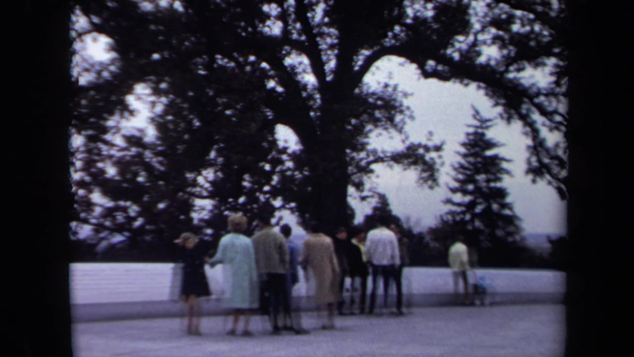 1970 Washington, DC home-movie frame showing tourists near a white memorial wall, with large trees and an overcast overlook beyond.