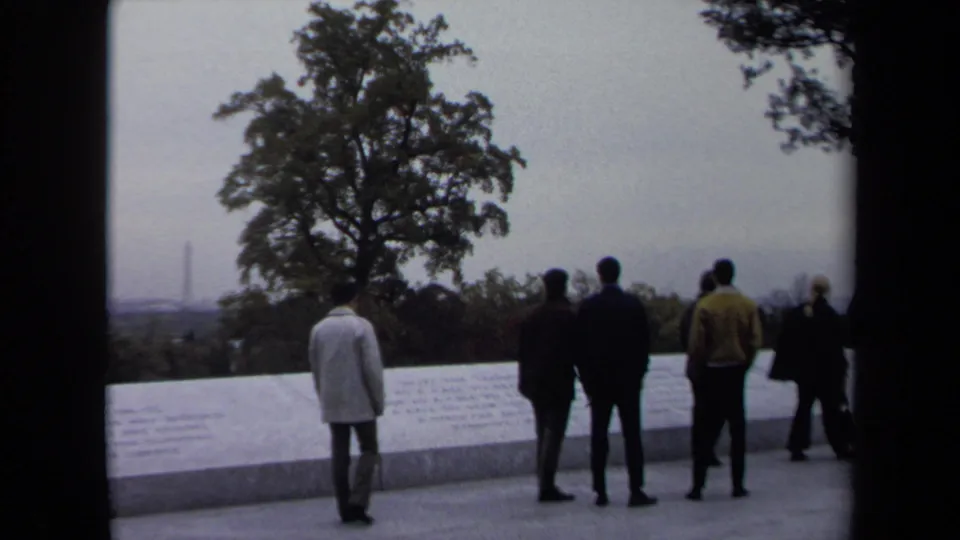 1970 Washington, DC frame showing visitors standing near a white memorial wall, with a tree, hazy skyline, and distant vertical monument-like form visible.