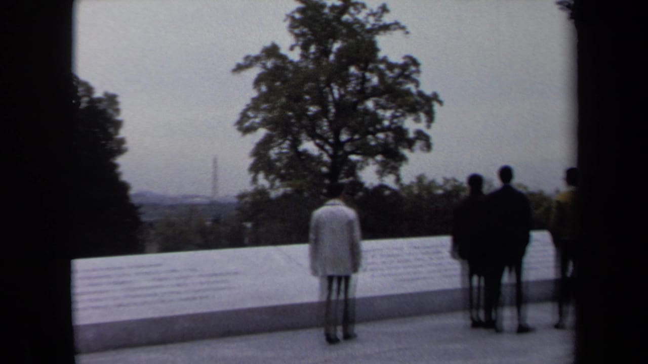 Visitors stand near an engraved stone wall with trees and the Washington Monument in the distance.