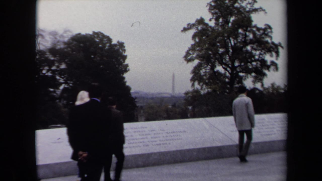 Washington DC visitors in coats move along a memorial wall while the Washington Monument appears in the hazy distance.
