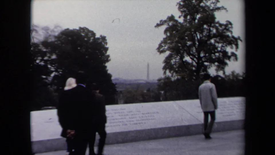Washington DC visitors in coats move along a memorial wall while the Washington Monument appears in the hazy distance.
