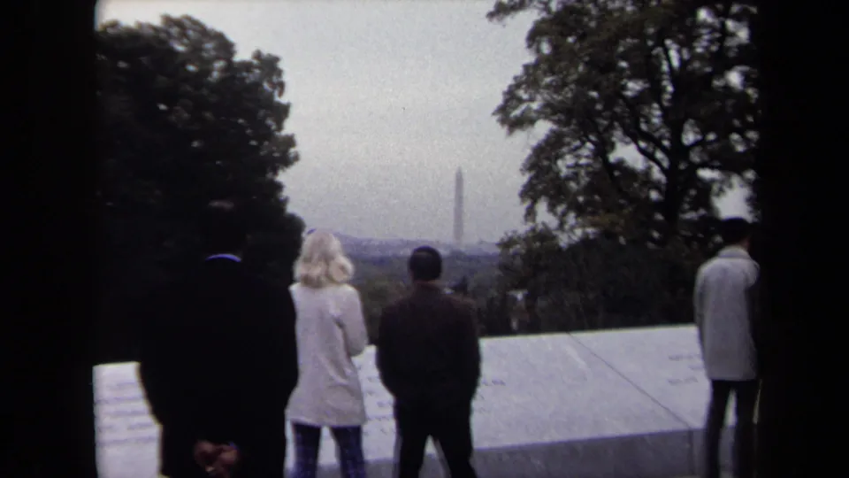 1970 home-movie frame of adults at a Washington DC memorial viewpoint, with a blonde woman and men facing the distant monument.