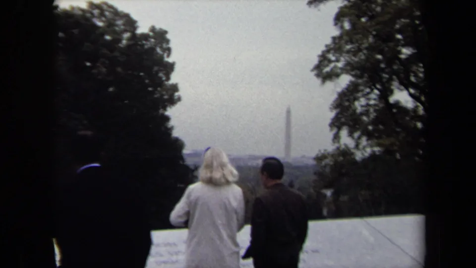 Visitors at a Washington DC memorial overlook face the distant Washington Monument, framed by trees and soft 8mm grain.