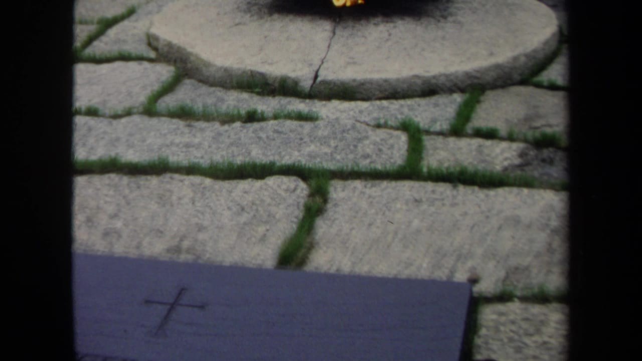 1970 Washington, DC frame showing part of a grave marker with a cross, with stone paving and grass seams in the background.