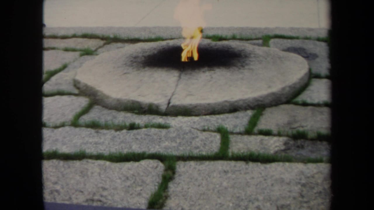 1970 8mm Washington, DC frame with a flame flickering on a round stone fire pit, bordered by gray pavers and patches of grass.