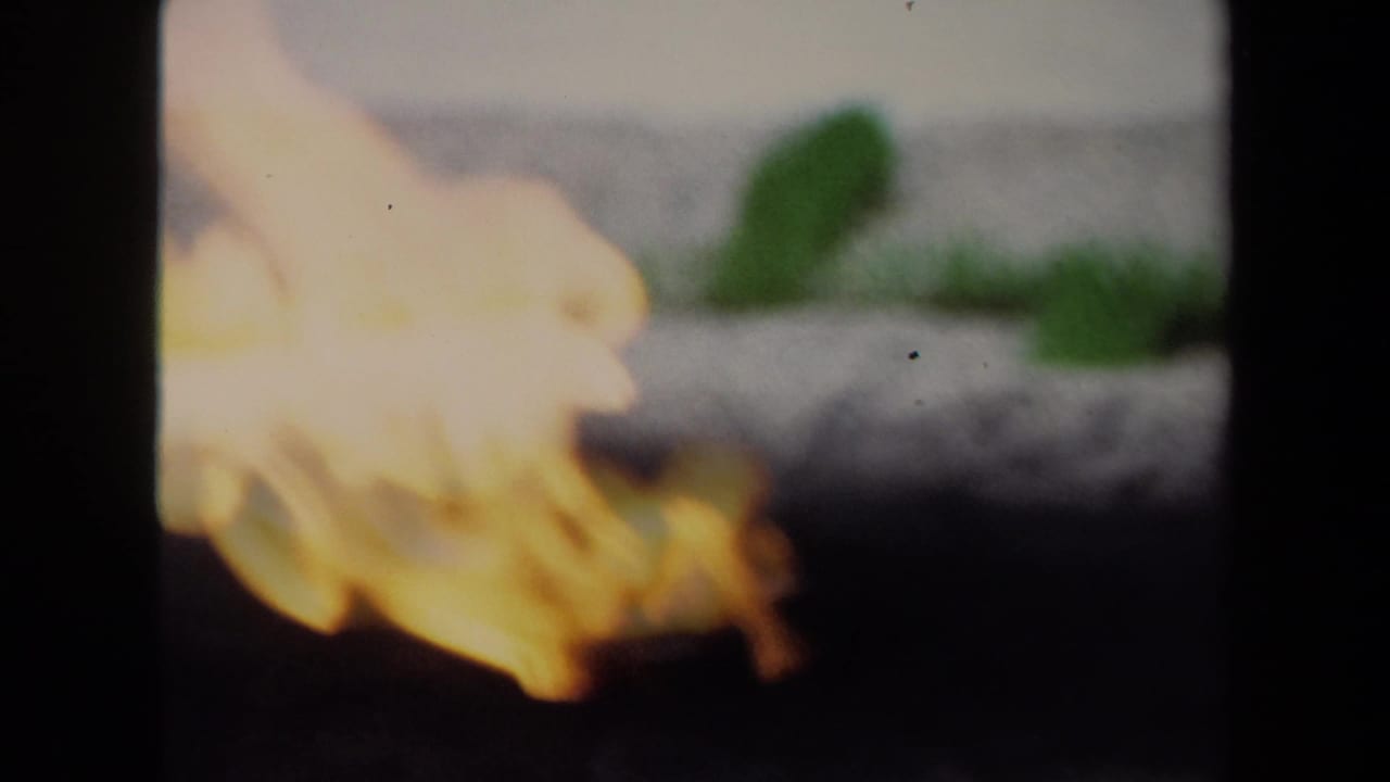 A close 1970 8mm frame shows warm flames in soft focus against a dark foreground, with film grain and heavy edge shadow.