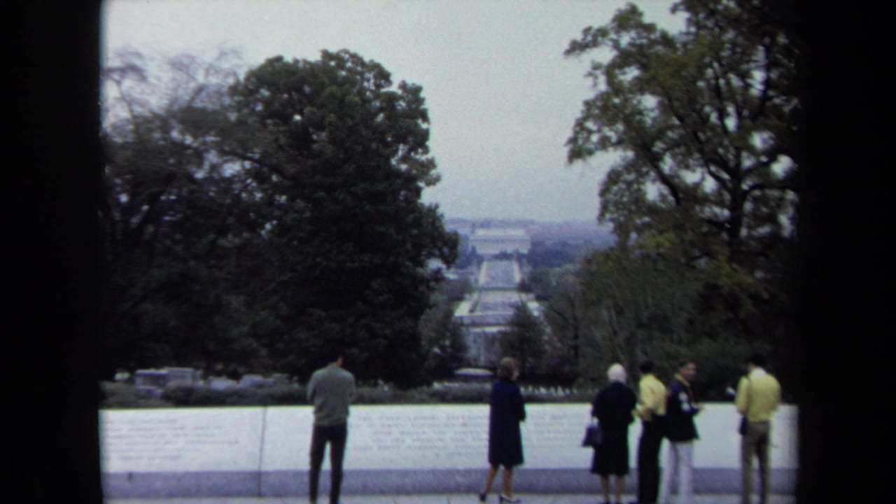 In Washington, DC, tourists stand along a pale stone overlook facing the Reflecting Pool axis and distant memorial architecture under an overcast sky.