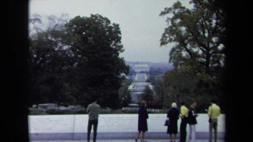 In Washington, DC, tourists stand along a pale stone overlook facing the Reflecting Pool axis and distant memorial architecture under an overcast sky.