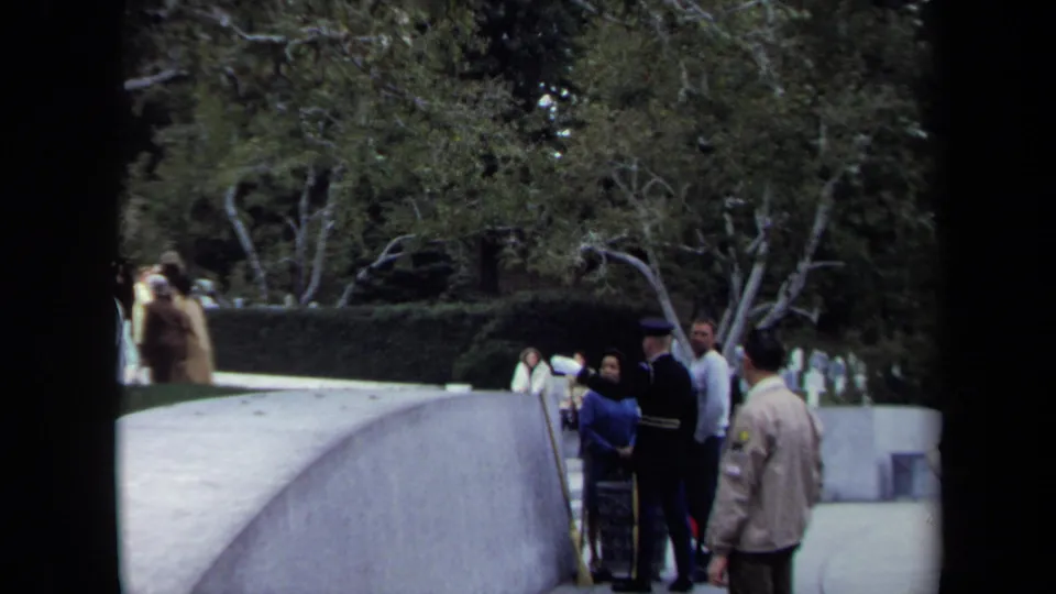 1970 Washington DC frame showing tourists gathered by a stone overlook near the memorial viewpoint, with trees and walkway behind.