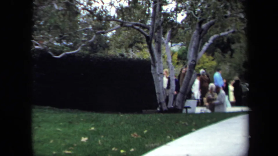 1970 Washington, DC park frame with visitors gathered near trees, a trimmed hedge, lawn, and a pale curved path.