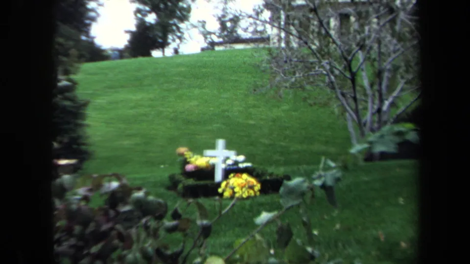1970 Washington, DC 8mm frame with a small white cross and flowers on a green cemetery-like garden slope.