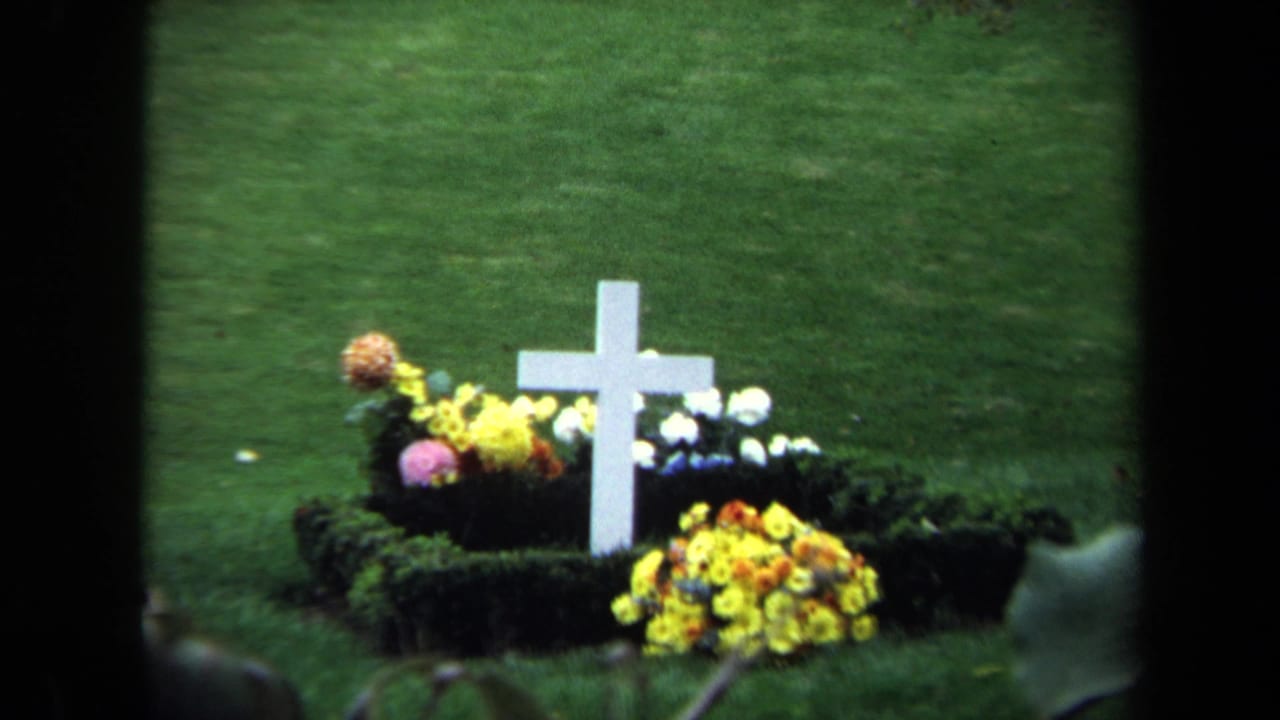 1970 Washington, DC 8mm frame showing a white cross grave marker surrounded by flowers on a grassy military cemetery plot.