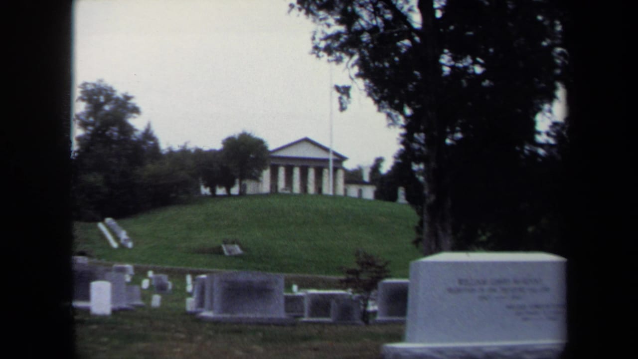 1970 8mm Washington, D.C. cemetery view showing a McAdoo gravestone on a grassy slope with trees and a classical building beyond.