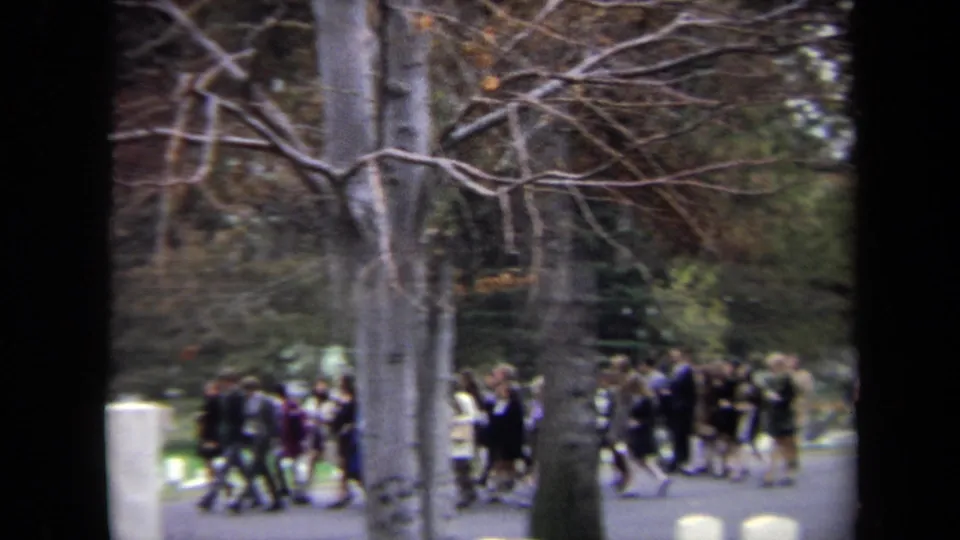 A handheld cemetery view shows groups of mourners or visitors walking along a path among headstones and bare trees.