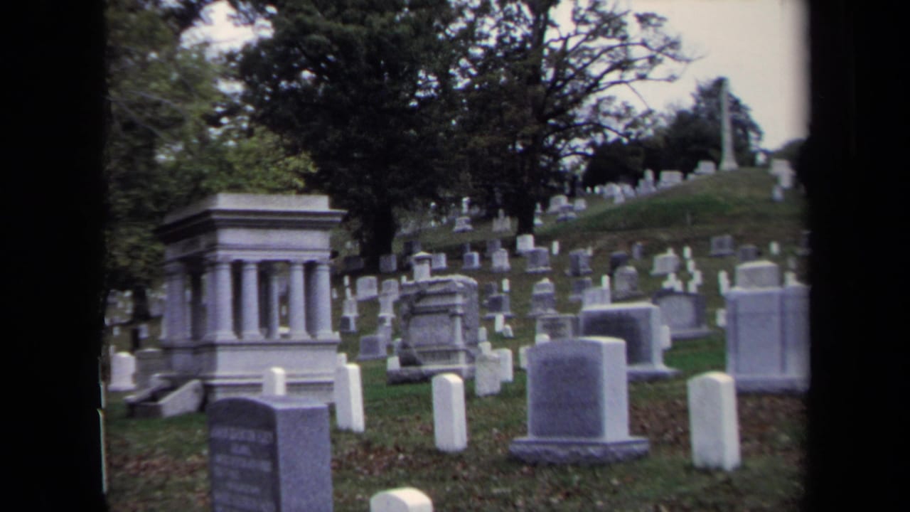 1970 Washington, DC 8mm frame showing a cemetery hillside with upright headstones, large monuments, grass, and mature trees.