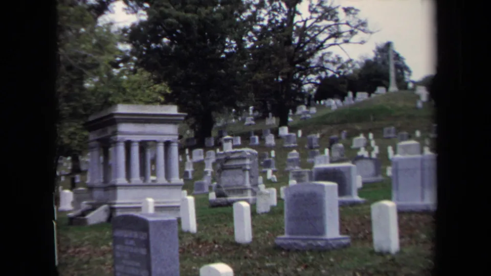 1970 Washington, DC 8mm frame showing a cemetery hillside with upright headstones, large monuments, grass, and mature trees.