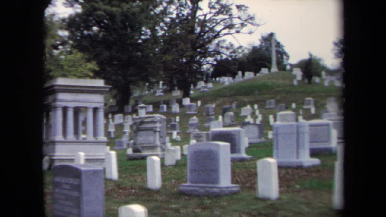 Washington, DC cemetery view from 1970 home-movie footage, showing rows of graves, a prominent monument, and trees on a hill.