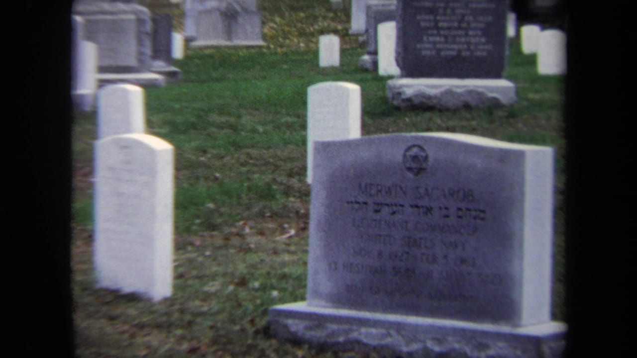 Closer cemetery view with a gray gravestone in the foreground and white markers behind it.