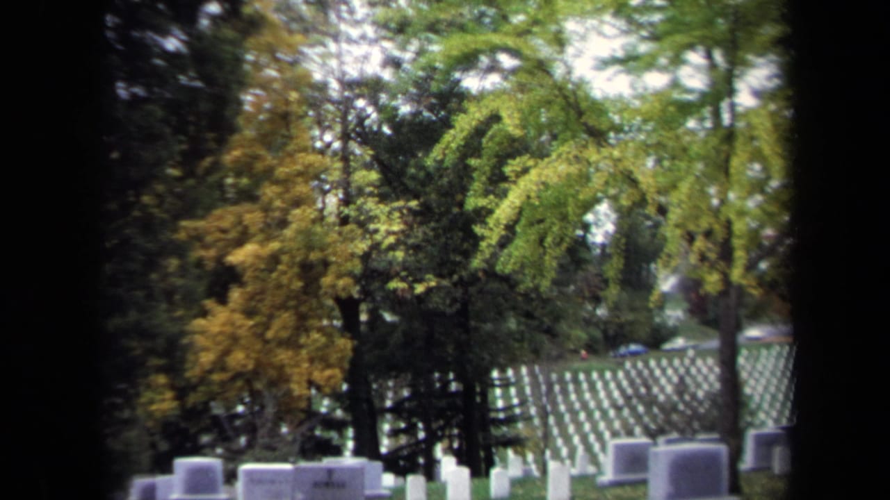 1970 Washington, DC home-movie frame showing white cemetery headstones on a grassy slope, seen through trees and dark film borders.