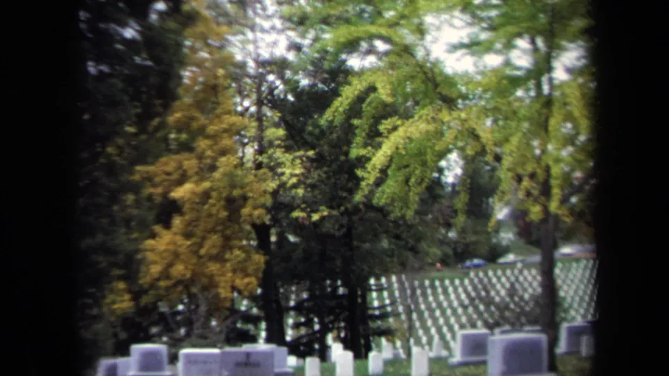 1970 Washington, DC home-movie frame showing white cemetery headstones on a grassy slope, seen through trees and dark film borders.