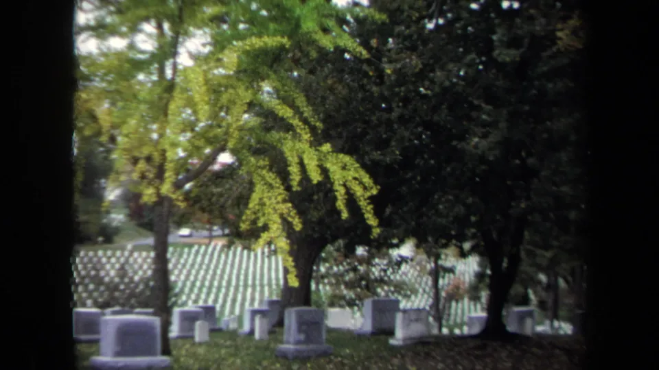 1970 Washington, DC 8mm frame with a wider cemetery view, showing white headstones beneath trees and a distant roadway.