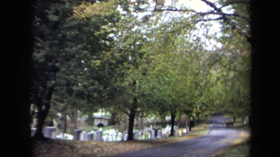 Washington, DC cemetery road curves through headstones and autumn foliage in a soft, grainy 1970 8mm frame.