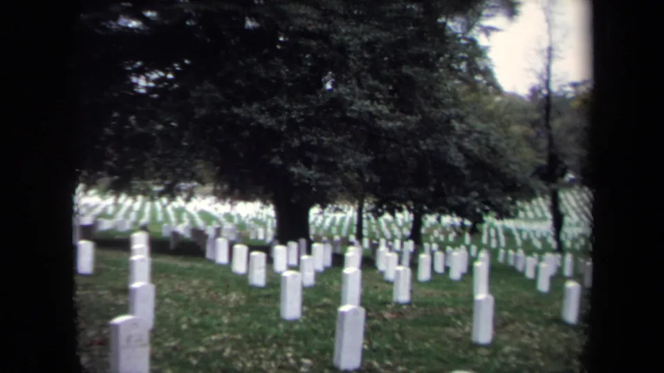 A handheld 8mm frame looks through trees toward white headstones on green grass in the Washington, DC cemetery setting.
