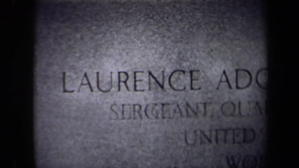 Home-movie frame showing the cropped tombstone inscription, including Laurence and sergeant text, with grain and handheld framing.
