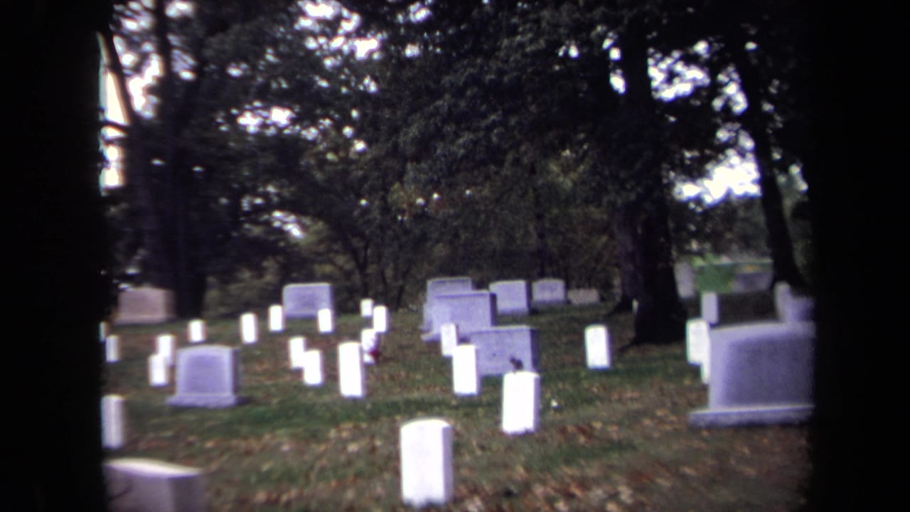 Rows of white headstones in a tree-shaded cemetery with dark film borders.
