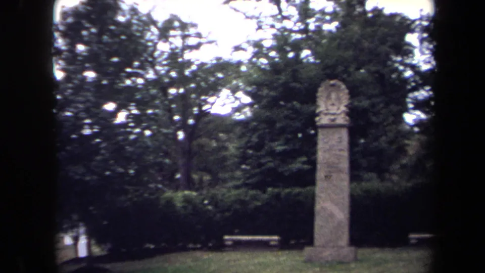 1970 8mm frame of a tall cemetery monolith in Washington, DC, with hedges, benches, trees, and dark film borders.