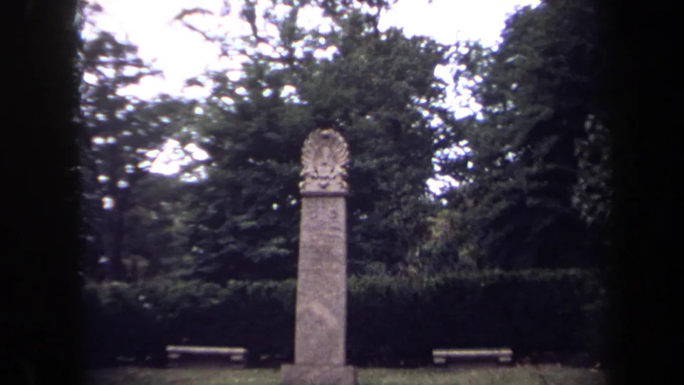 A tall stone monument in a Washington, DC cemetery appears between dark 8mm frame edges, with benches and dense greenery behind it.