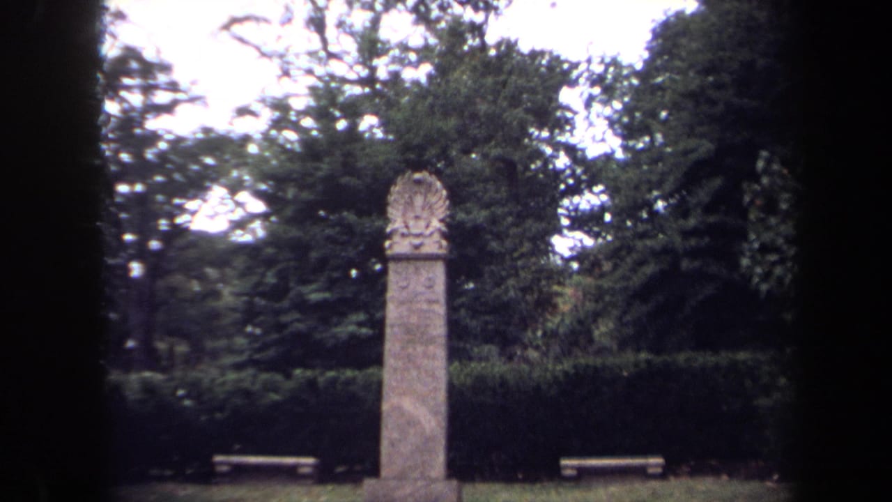 1970 Washington, DC home-movie frame showing a tall stone park monument with carved crest, hedge, trees, and benches.