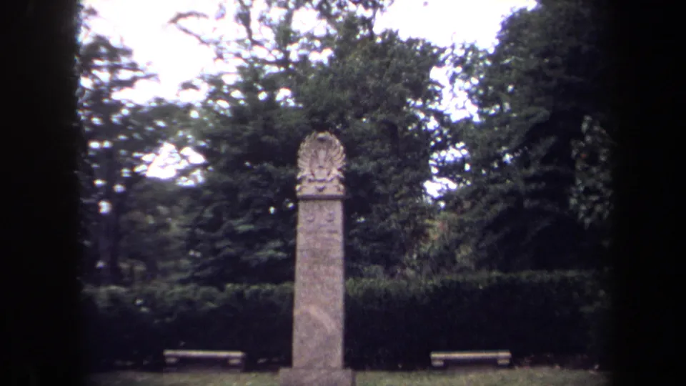 1970 Washington, DC home-movie frame showing a tall stone park monument with carved crest, hedge, trees, and benches.