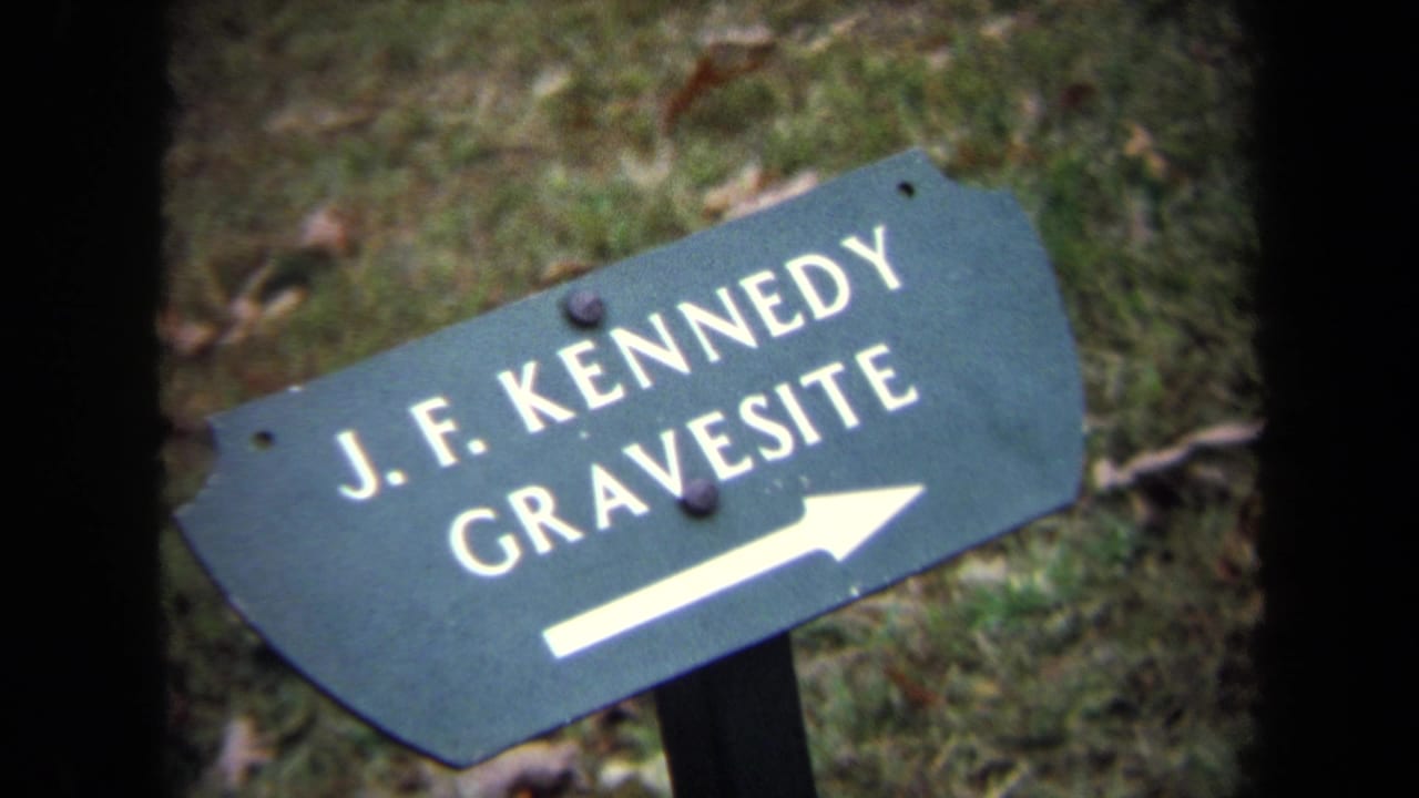Green Arlington cemetery directional sign reading J.F. Kennedy Gravesite, with a white arrow pointing right over grass and fallen leaves.